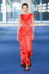 Model walking a fashion runway wearing a sleeveless red satin jumpsuit with wide legs on a blue catwalk.