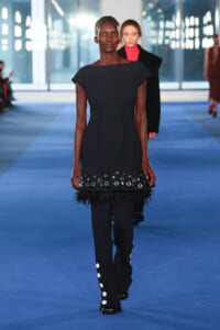 Model walks a blue runway in a dark, fitted dress with metallic eyelets and feather trim at the hem, paired with matching pants and boots. Behind her, another model is seen in red and black.