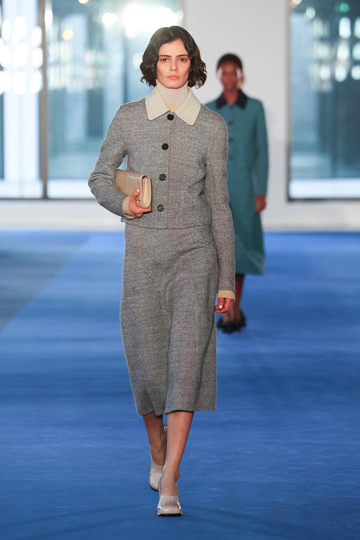 Model in a gray textured suit with a cream collar walks the blue runway, holding a beige clutch.