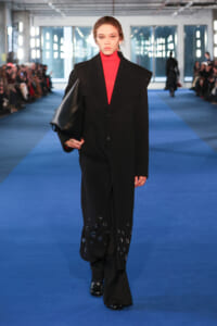 Model walking a blue runway in a black oversized coat over a red turtleneck, carrying a black clutch.