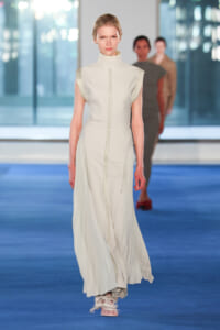 Model walking on a blue runway wearing a cream sleeveless high-neck gown with front seam and pleated panels at the hips, paired with fringe sandals.