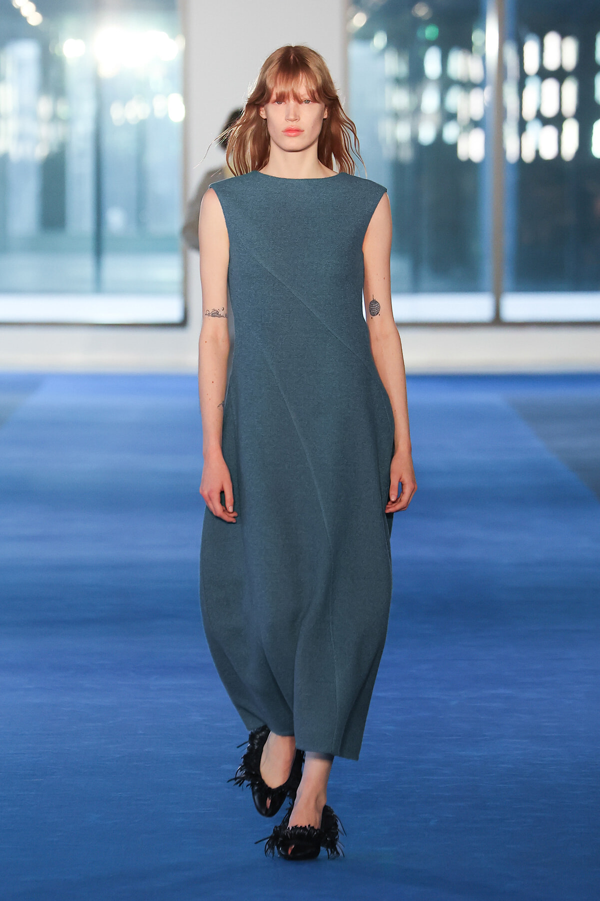 Model striding down a blue runway in a sleeveless teal dress with a diagonal seam and loose silhouette, paired with black feathered flats