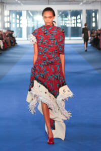 Model walks a fashion runway in a burgundy dress with teal leaf print and large white fringed hem, red shoes visible as she strides toward the camera on a blue catwalk.