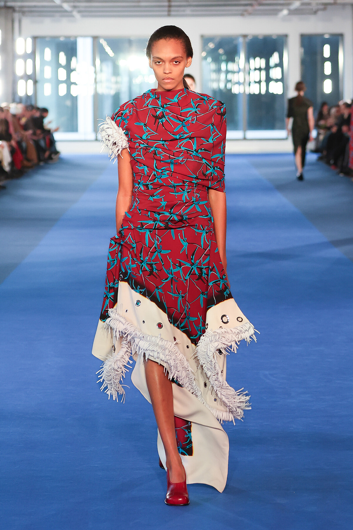 Model walks a fashion runway in a burgundy dress with teal leaf print and large white fringed hem, red shoes visible as she strides toward the camera on a blue catwalk.