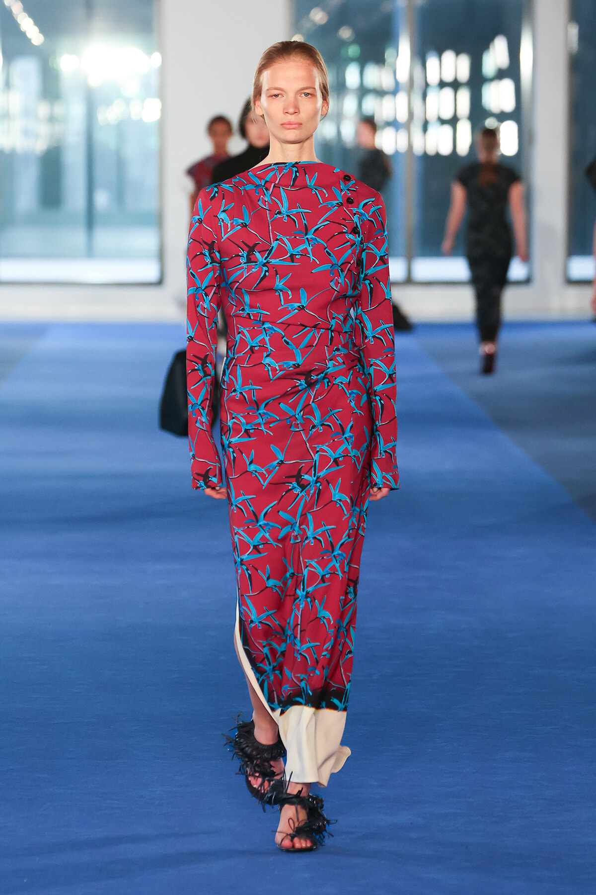 Model wearing a burgundy long-sleeve dress with a blue bamboo print on a blue runway, paired with feathered black sandals.