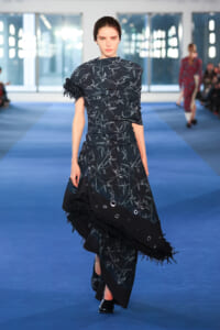Female model on a blue runway wearing a navy floral evening gown with fringe and metal grommet trim.