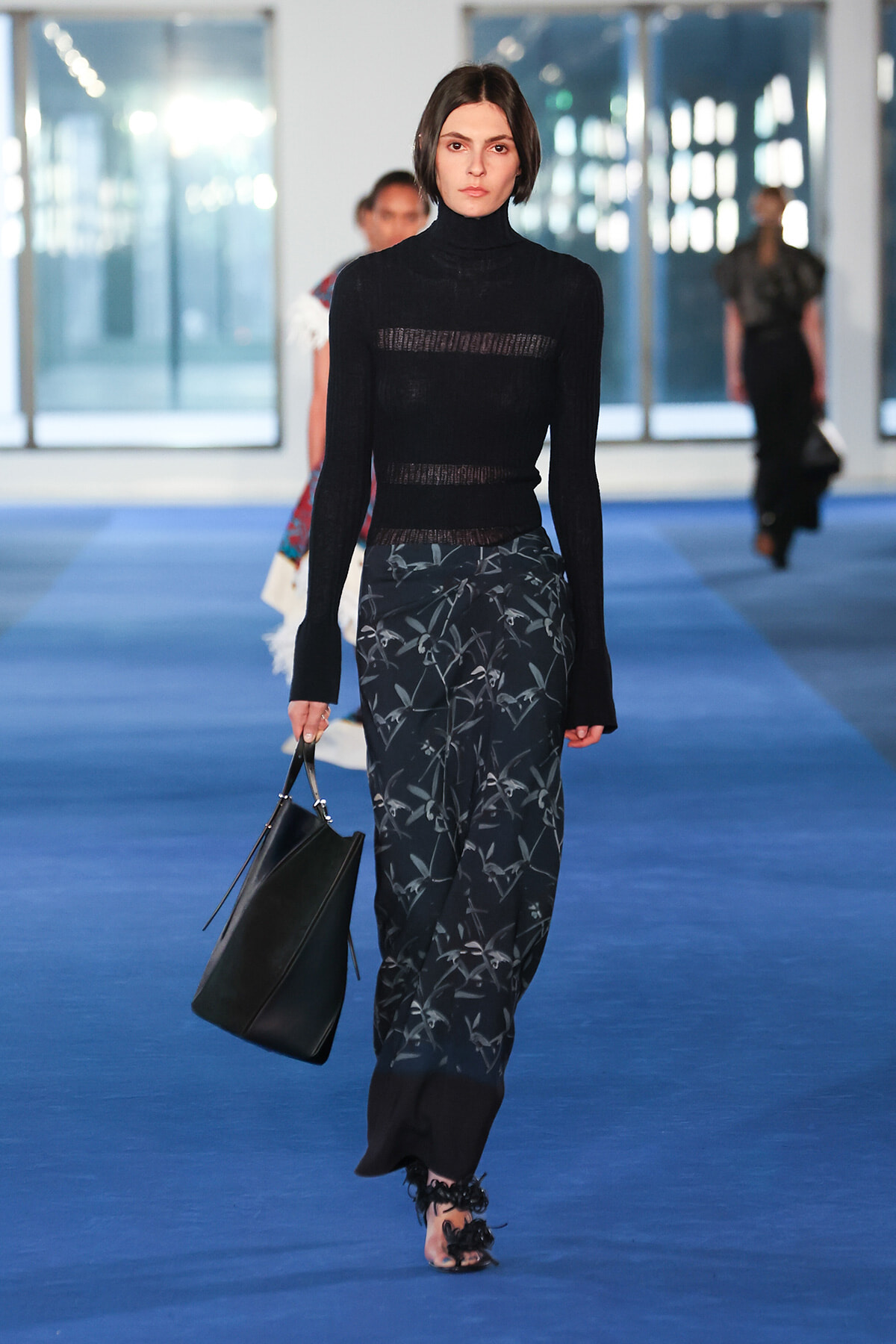 Model walking a runway in a black sheer turtleneck top and dark floral-patterned wide-leg pants, carrying a black handbag on a blue catwalk.
