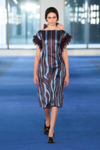 Model walking the runway in a blue-and-maroon striped knee-length dress with ruffled fringe sleeves.