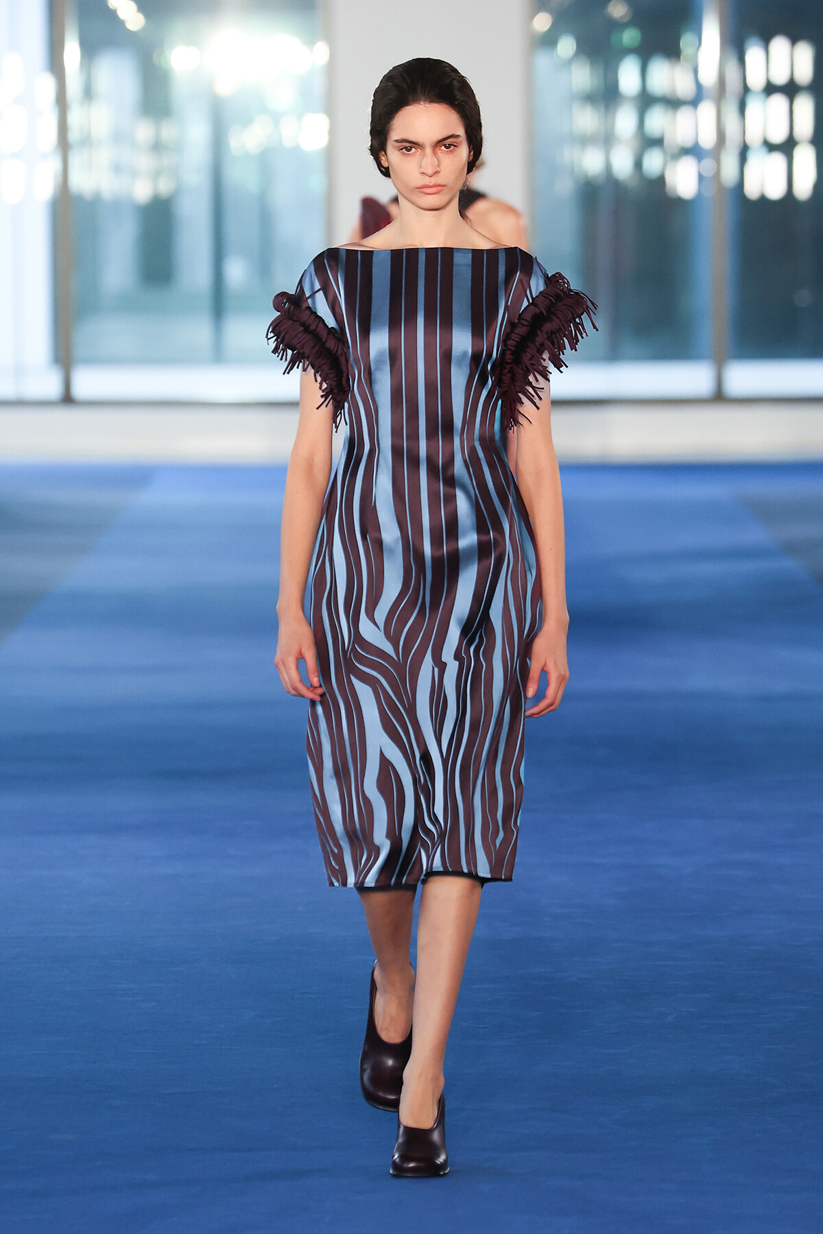 Model walking the runway in a blue-and-maroon striped knee-length dress with ruffled fringe sleeves.