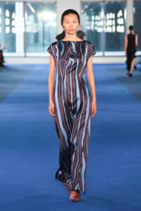 Model walking a blue-and-brown vertical-striped jumpsuit on a fashion runway in a bright venue