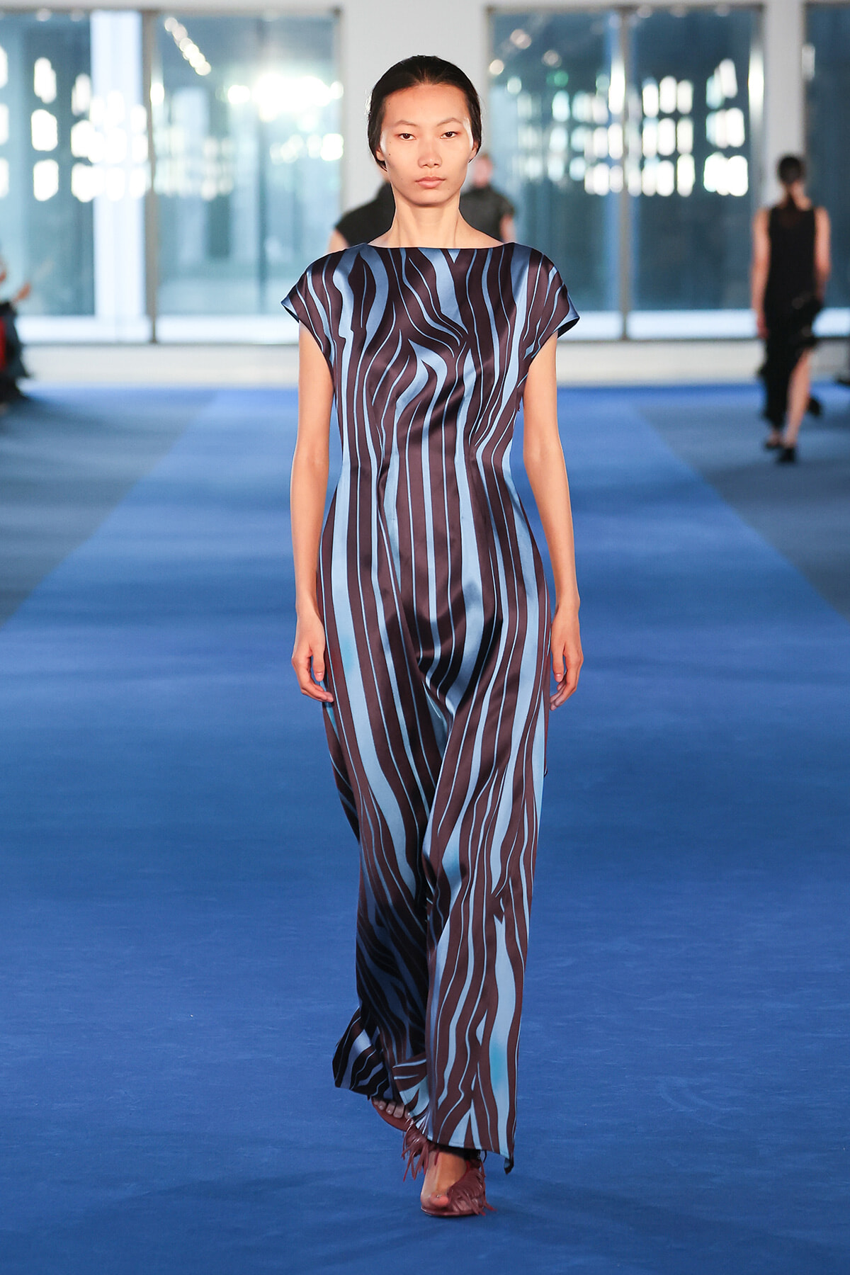 Model walking a blue-and-brown vertical-striped jumpsuit on a fashion runway in a bright venue