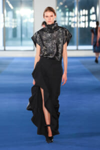 Model on a runway wearing a black high-neck satin top with a dragonfly print and a voluminous, ruffled skirt with a thigh-high slit.