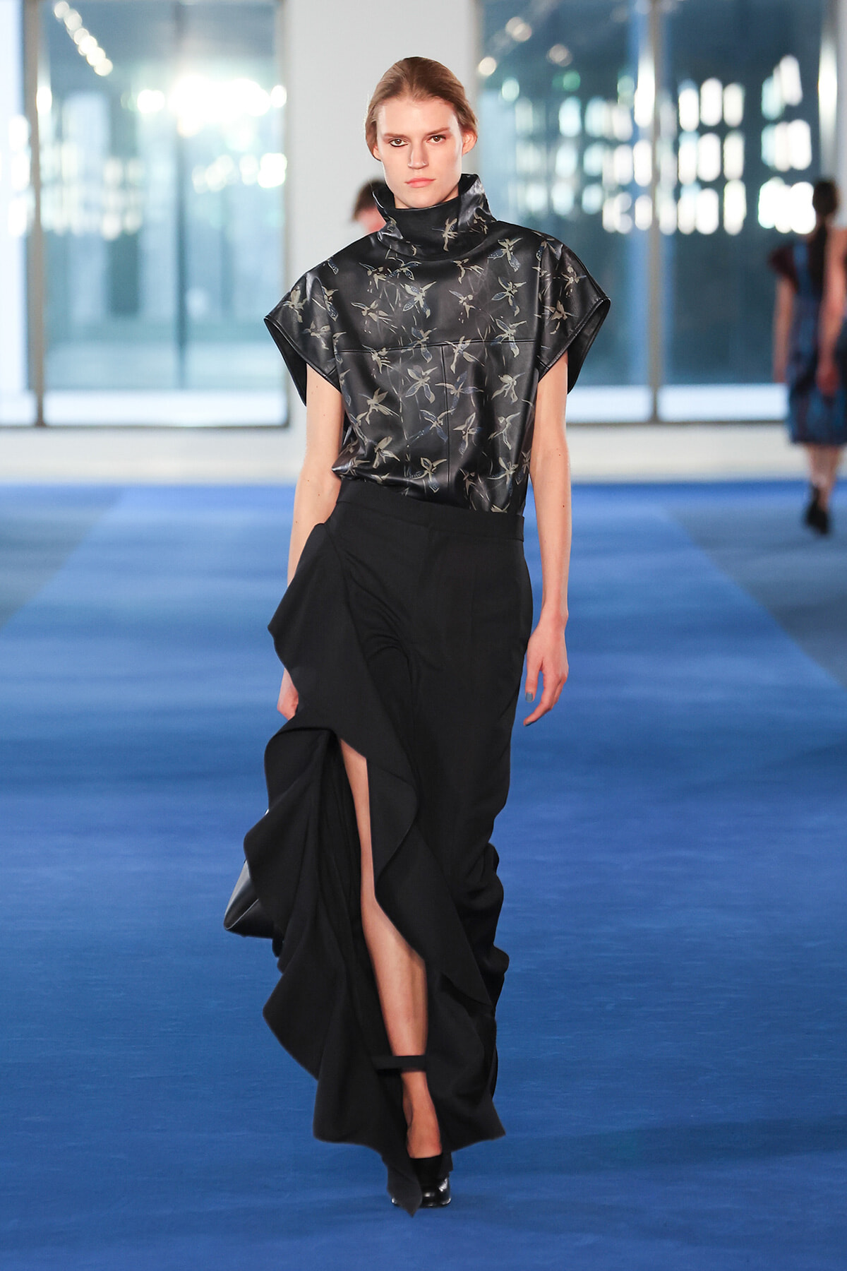 Model on a runway wearing a black high-neck satin top with a dragonfly print and a voluminous, ruffled skirt with a thigh-high slit.