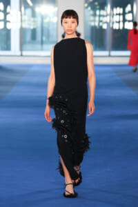 Model walking a blue runway in a sleeveless black gown with fringe and silver eyelet detailing along the hip area.