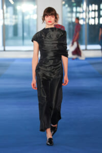 Model walking a blue runway in a black satin, draped, one-shoulder dress with short brown hair and black shoes.