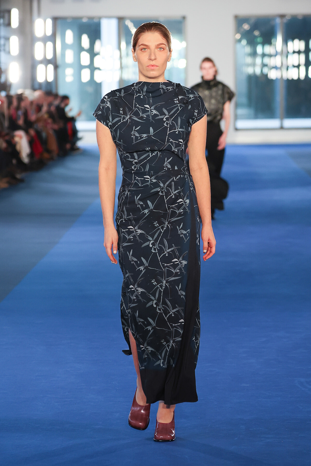 Model walks down a blue runway in a dark blue floral dress with a side slit and burgundy shoes; audience visible on the left.