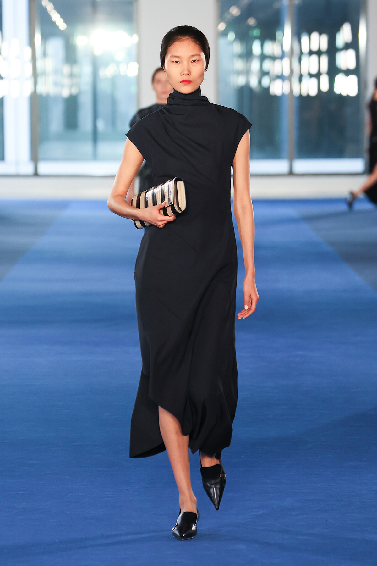 Model walking on a blue runway in a black high-neck dress, holding a striped clutch.