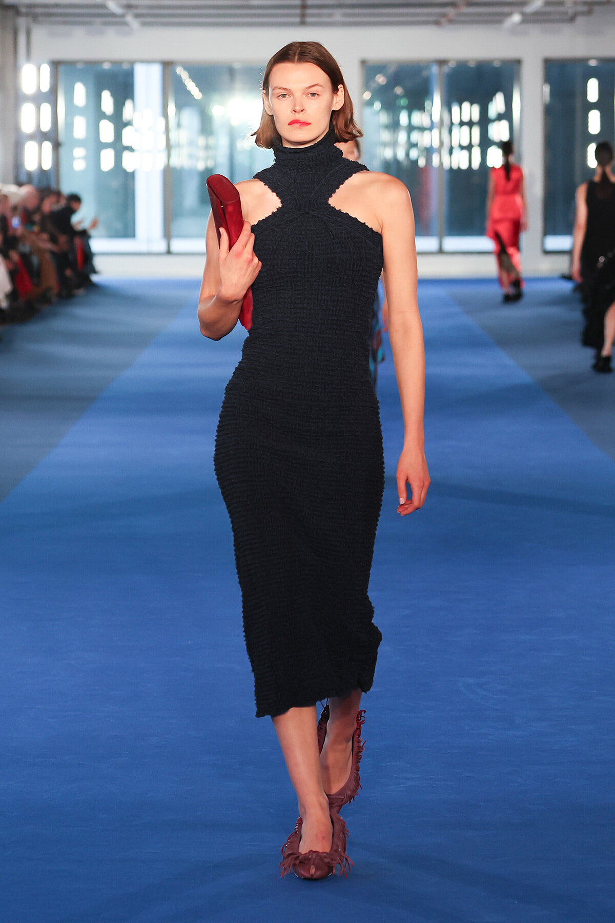 Model walking a blue runway in a black textured halter dress with cutouts at the shoulders, carrying a red clutch.