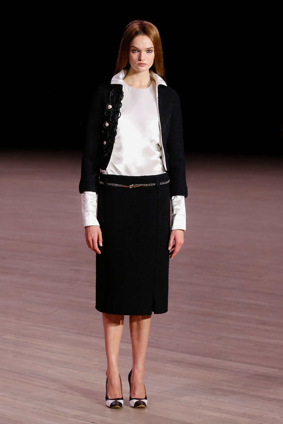 Model on a runway wearing a black two-piece suit with a white blouse and cuffs, belt, and black-and-white heels against a dark background.