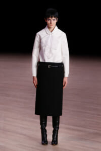 Model on a runway wearing a white blouse with a draped front, a black midi skirt, a belt, and knee-high black boots.