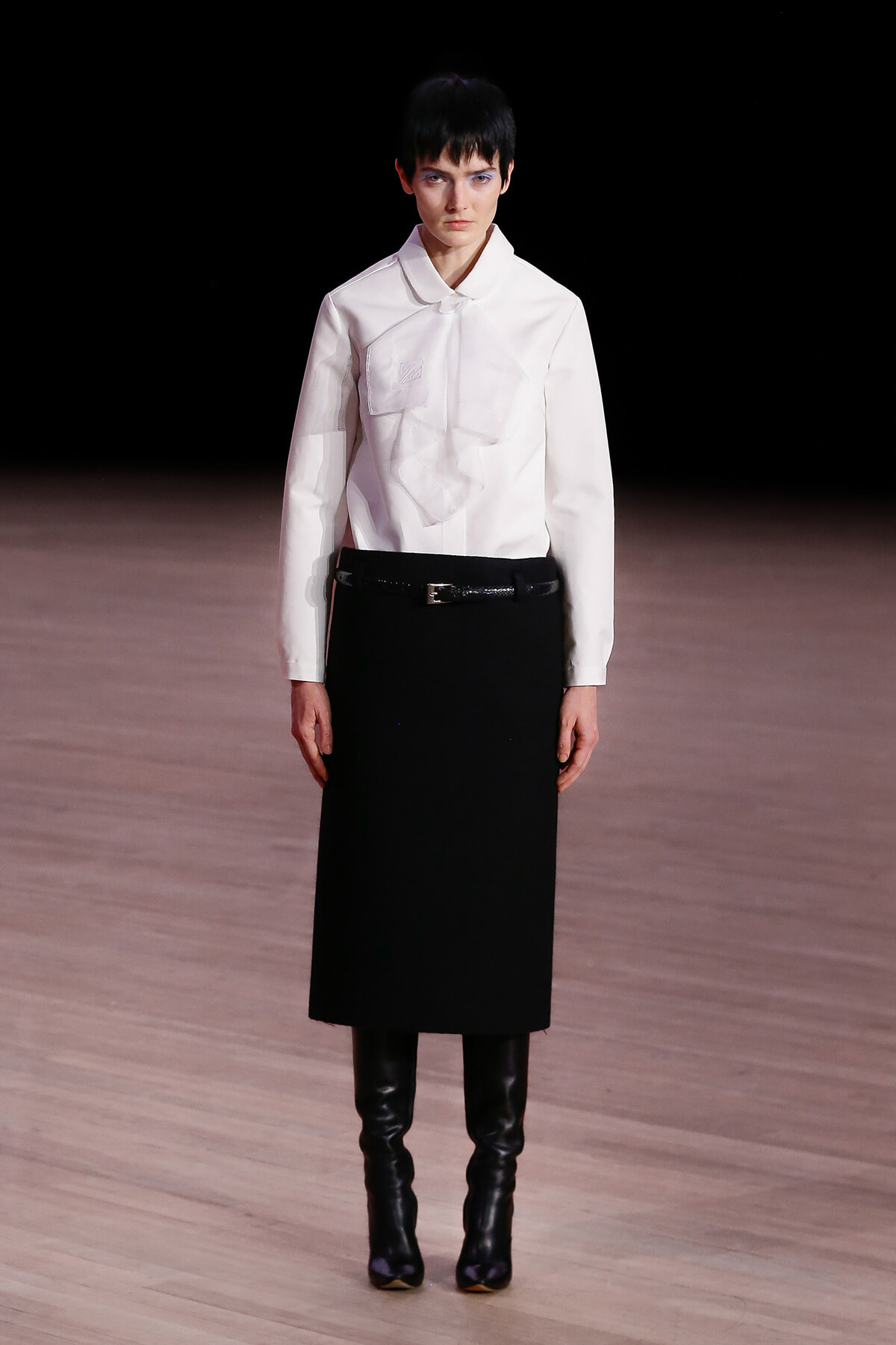 Model on a runway wearing a white blouse with a draped front, a black midi skirt, a belt, and knee-high black boots.