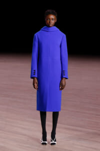 Model walking a runway in a royal blue knee-length coat with a high collar, paired with black tights and white-strap heels, against a dark background.
