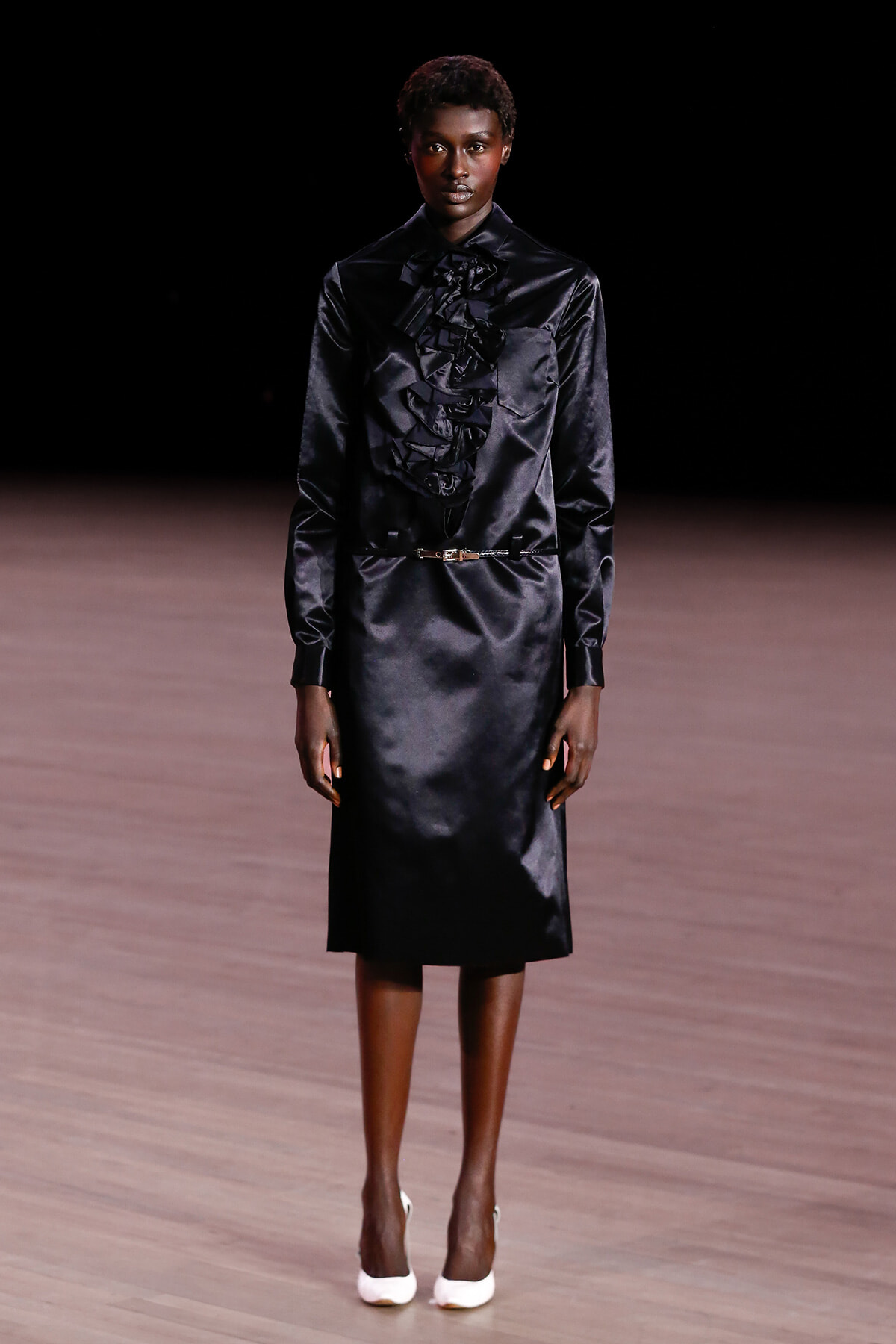 Model wearing a black satin blouse with a ruffled front tucked into a black midi skirt on a runway, paired with white heels.