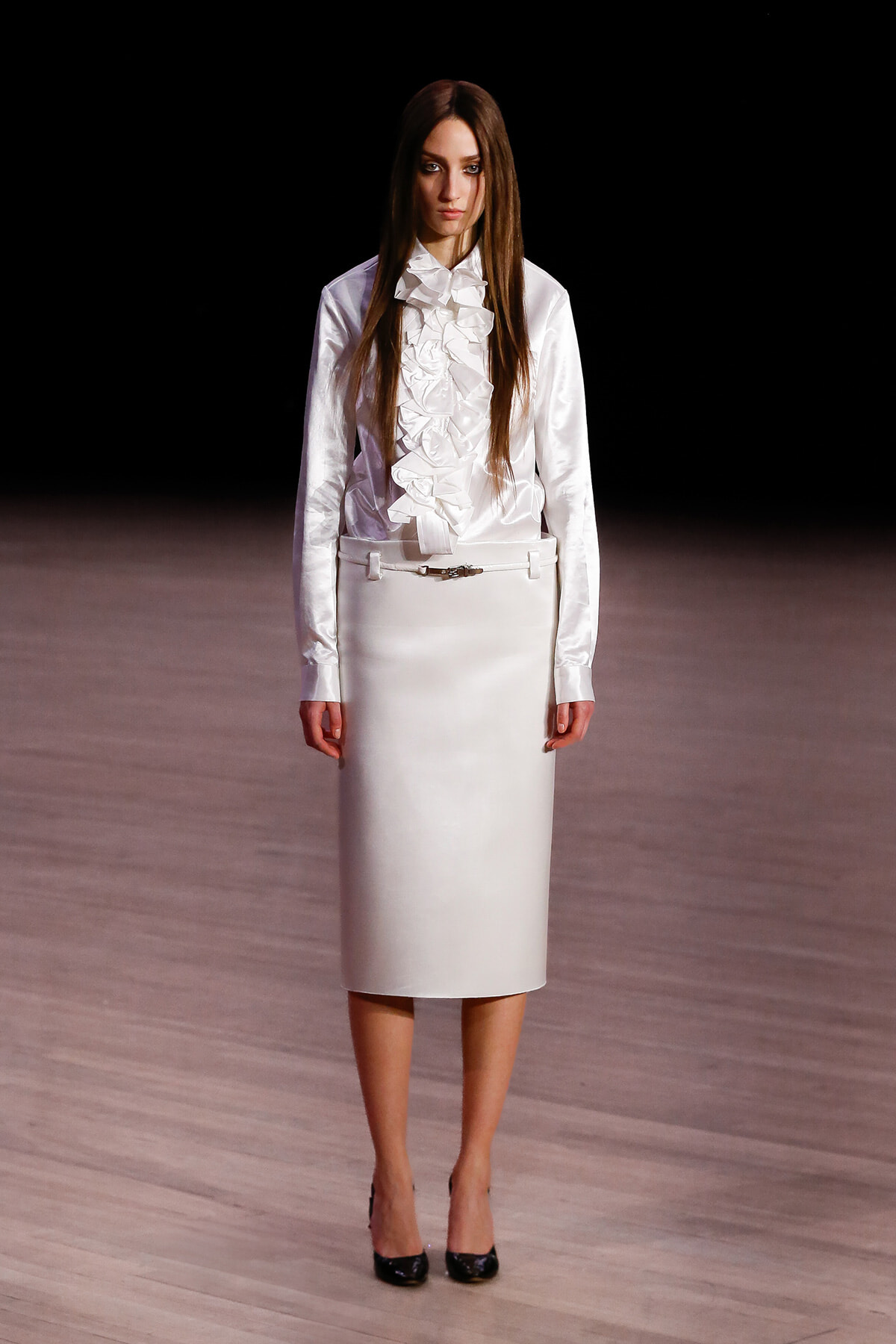 Female model on a runway wearing a white satin blouse with a ruffled front and a white knee-length pencil skirt, black heels.