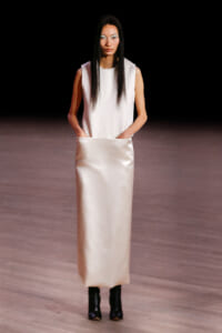 Model walks a runway in a sleeveless white top and long satin cream skirt with pockets, wearing black ankle boots.