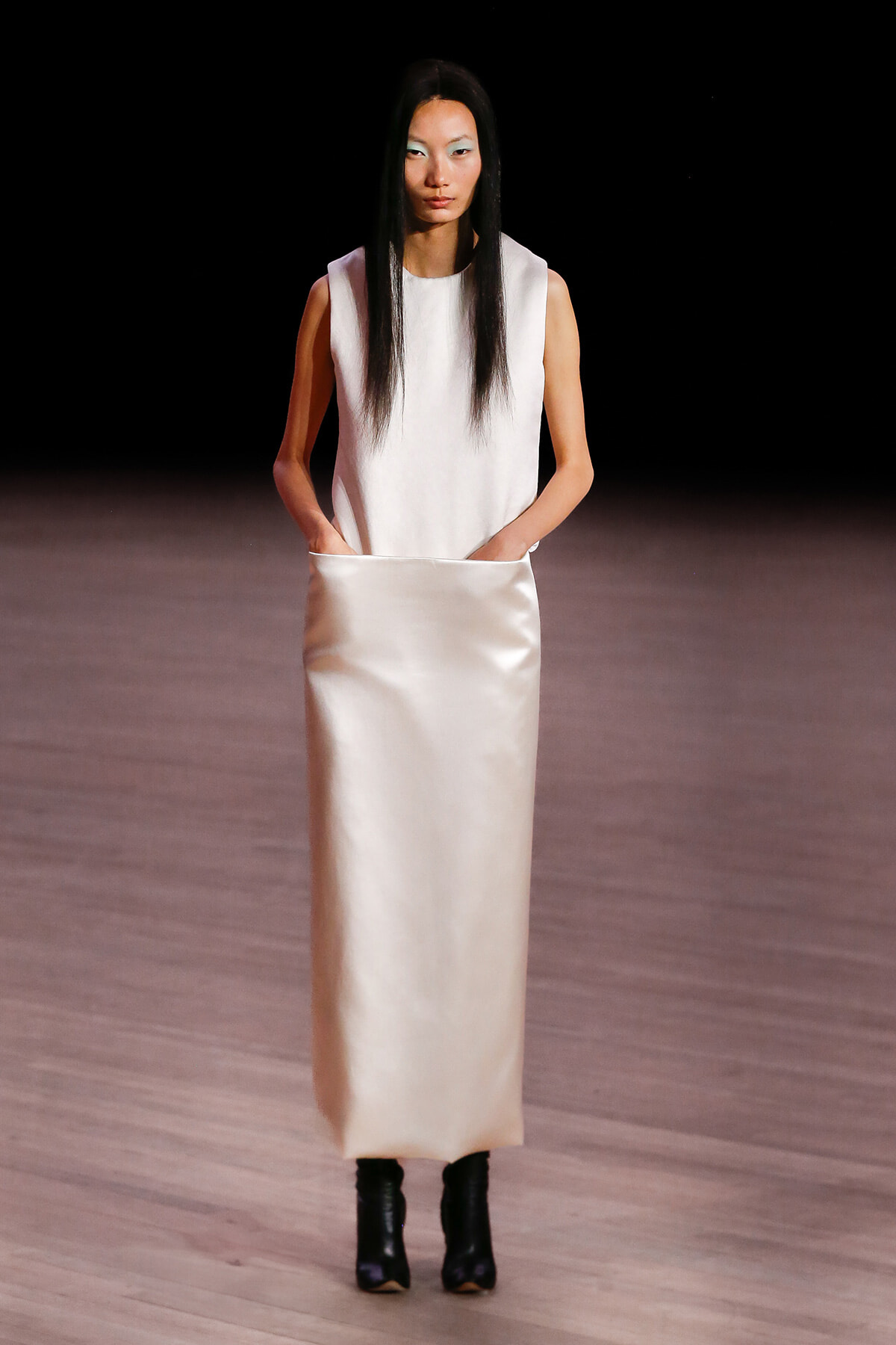 Model walks a runway in a sleeveless white top and long satin cream skirt with pockets, wearing black ankle boots.