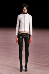 Model on a runway wearing a white zip-front jacket, black leather shorts with a belt, sheer black tights, and white-strap heels.