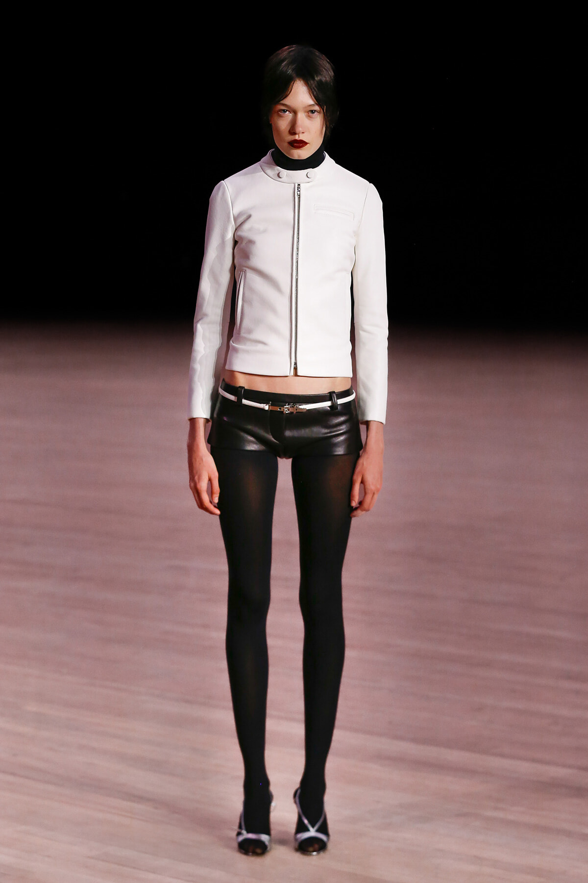 Model on a runway wearing a white zip-front jacket, black leather shorts with a belt, sheer black tights, and white-strap heels.