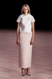 Model on a runway wearing a white short-sleeve top with a pale pink floral-embroidered maxi skirt.