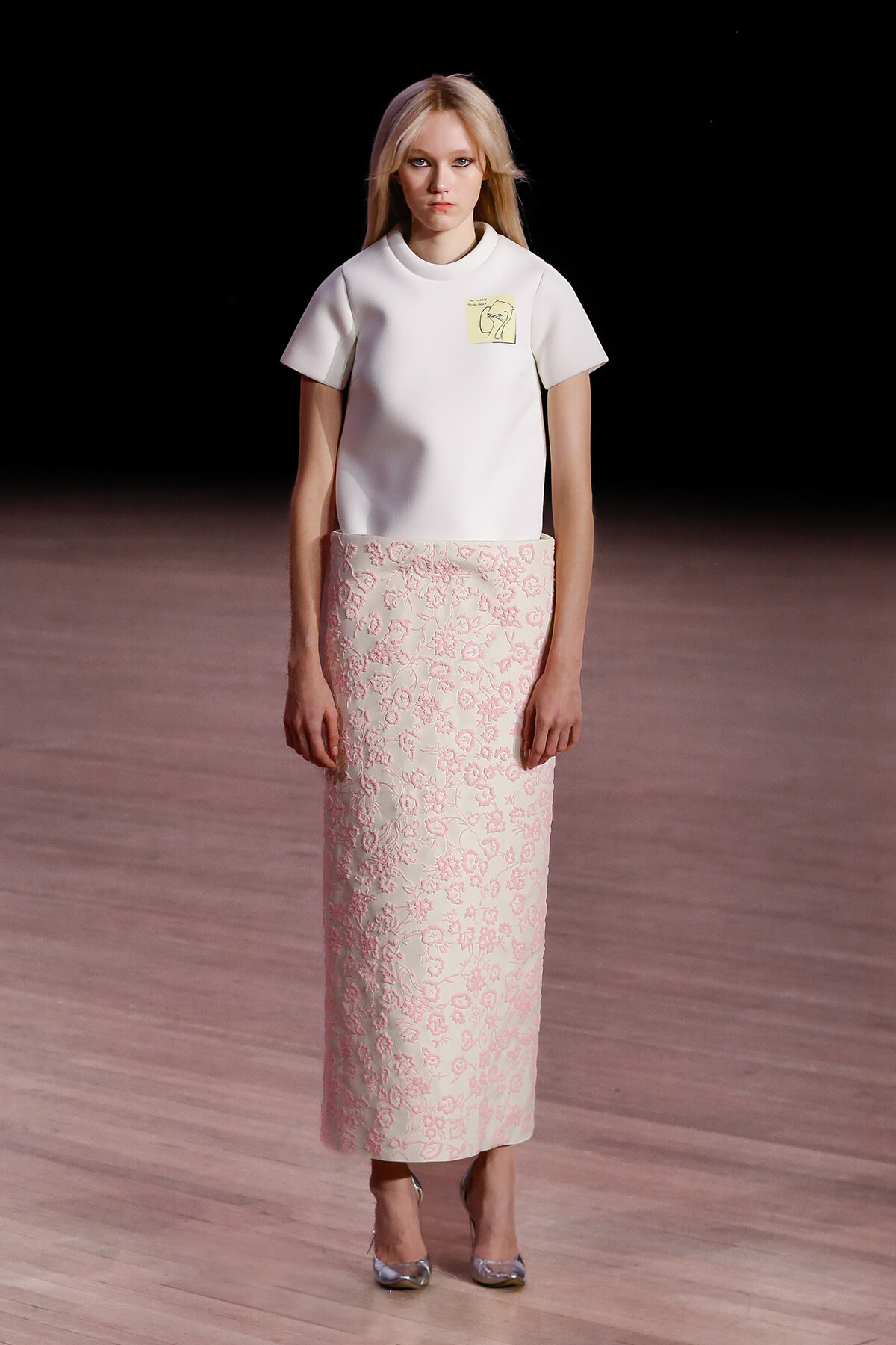 Model on a runway wearing a white short-sleeve top with a pale pink floral-embroidered maxi skirt.