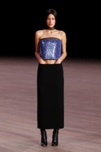 Model wearing a blue sequined strapless top and a long black skirt on a runway stage.