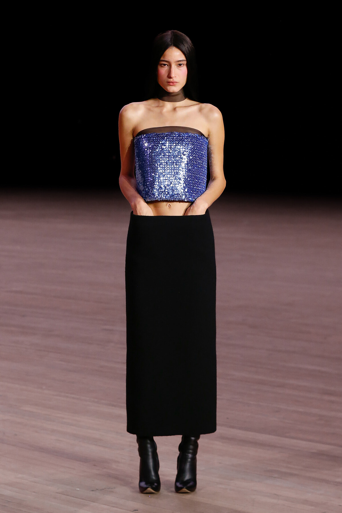 Model wearing a blue sequined strapless top and a long black skirt on a runway stage.