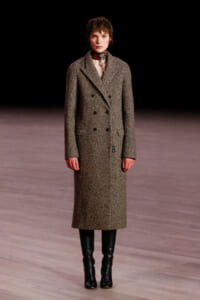 Runway model wearing a knee-length brown tweed double-breasted coat with black knee-high boots.