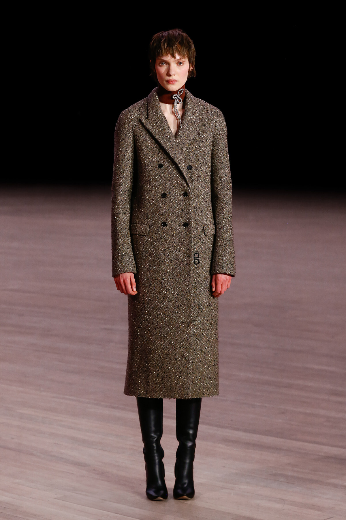 Runway model wearing a knee-length brown tweed double-breasted coat with black knee-high boots.