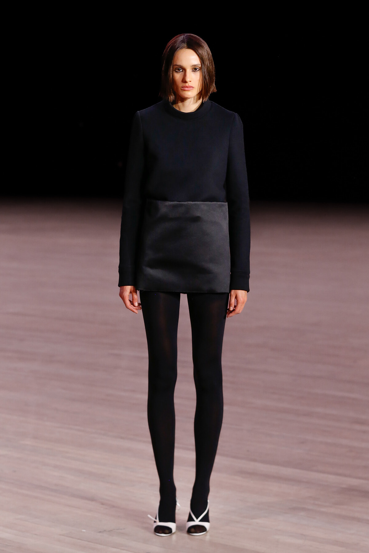 Model walks the runway in a black long-sleeve top with a shiny dark gray waist panel, pairing it with black tights and white-heeled sandals.