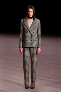 Model on a runway wearing a gray checkered double-breasted suit with matching trousers and black shoes.