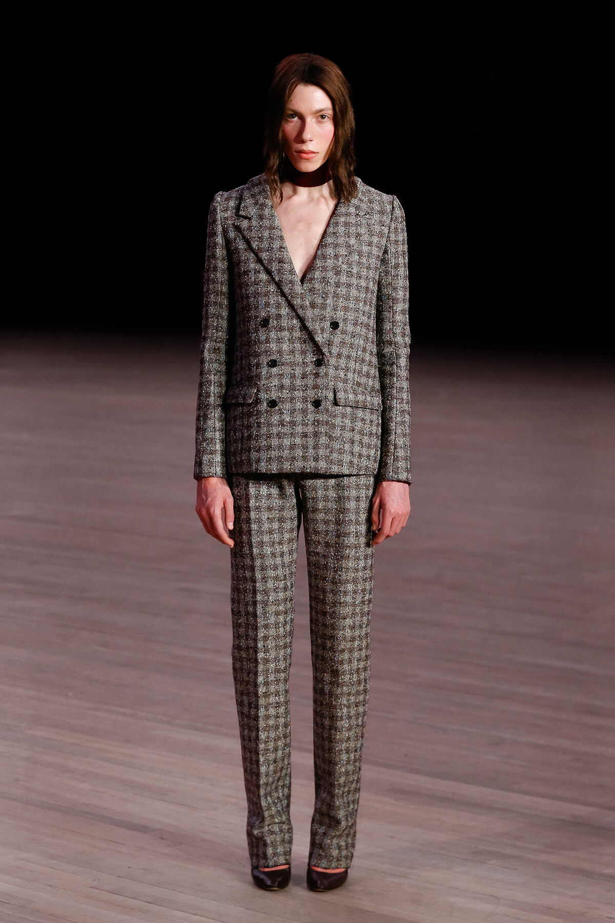 Model on a runway wearing a gray checkered double-breasted suit with matching trousers and black shoes.