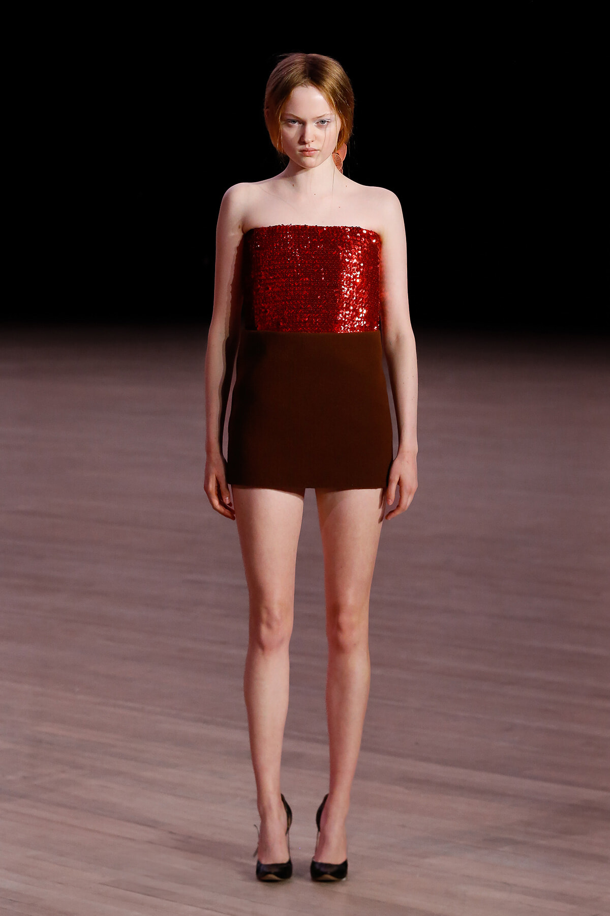 Model on runway wearing a strapless red sequin top and dark brown mini skirt, with black heels.
