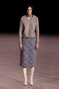 Model walking a fashion show runway in a beige knit sweater and a plaid midi skirt with a thin red belt, paired with cream knee-high boots against a dark backdrop.