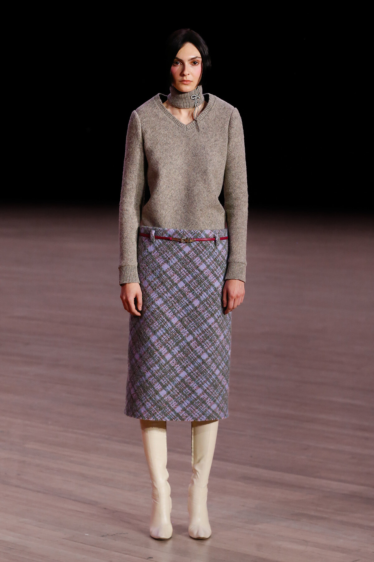 Model walking a fashion show runway in a beige knit sweater and a plaid midi skirt with a thin red belt, paired with cream knee-high boots against a dark backdrop.