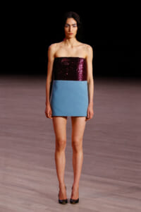 Model walking a runway in a strapless color-block mini dress: burgundy sequin top and light blue skirt, paired with black heels.