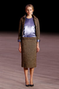 Model on a runway wearing a brown tweed jacket and matching midi skirt with a blue satin blouse and black heels.