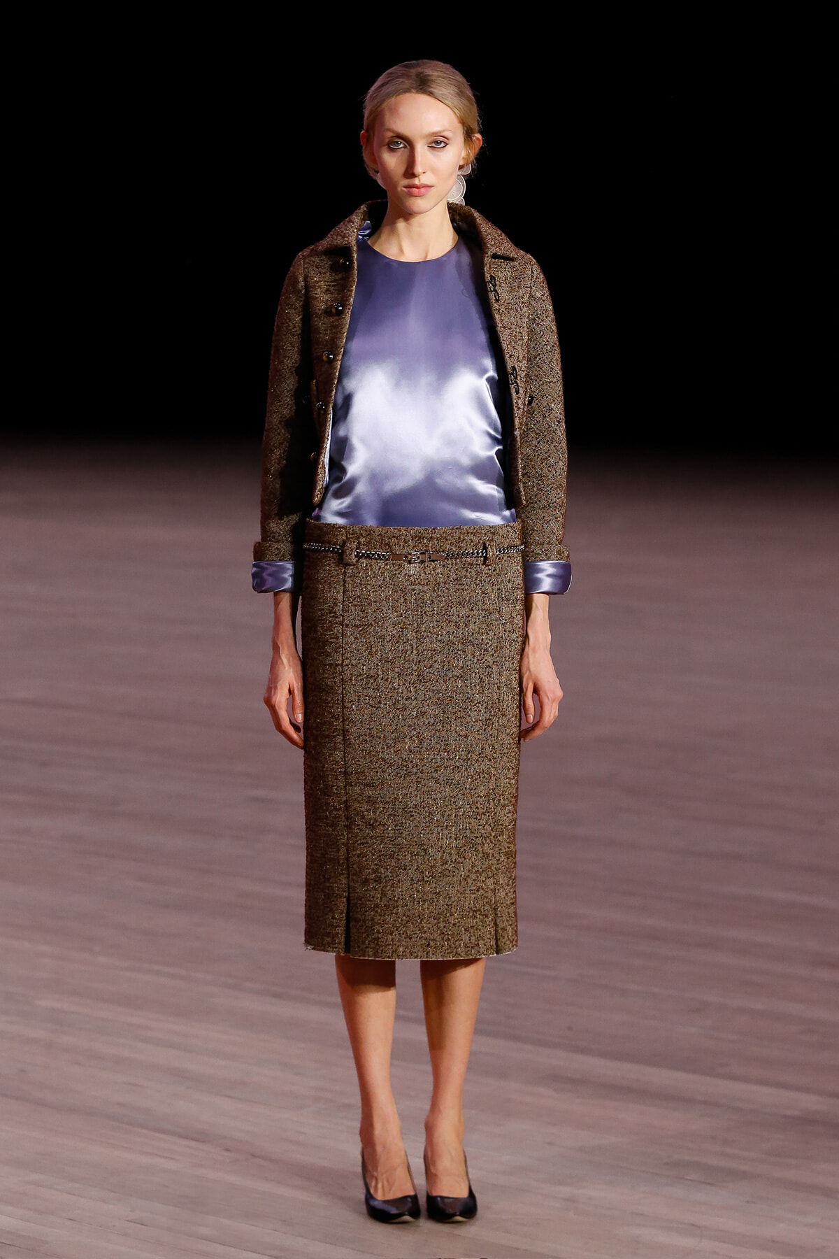 Model on a runway wearing a brown tweed jacket and matching midi skirt with a blue satin blouse and black heels.
