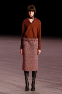 Female model walking a runway in a rust V-neck sweater, pink tweed knee-length skirt with a belt, and black knee-high boots, with a silver scarf tied at the neck.