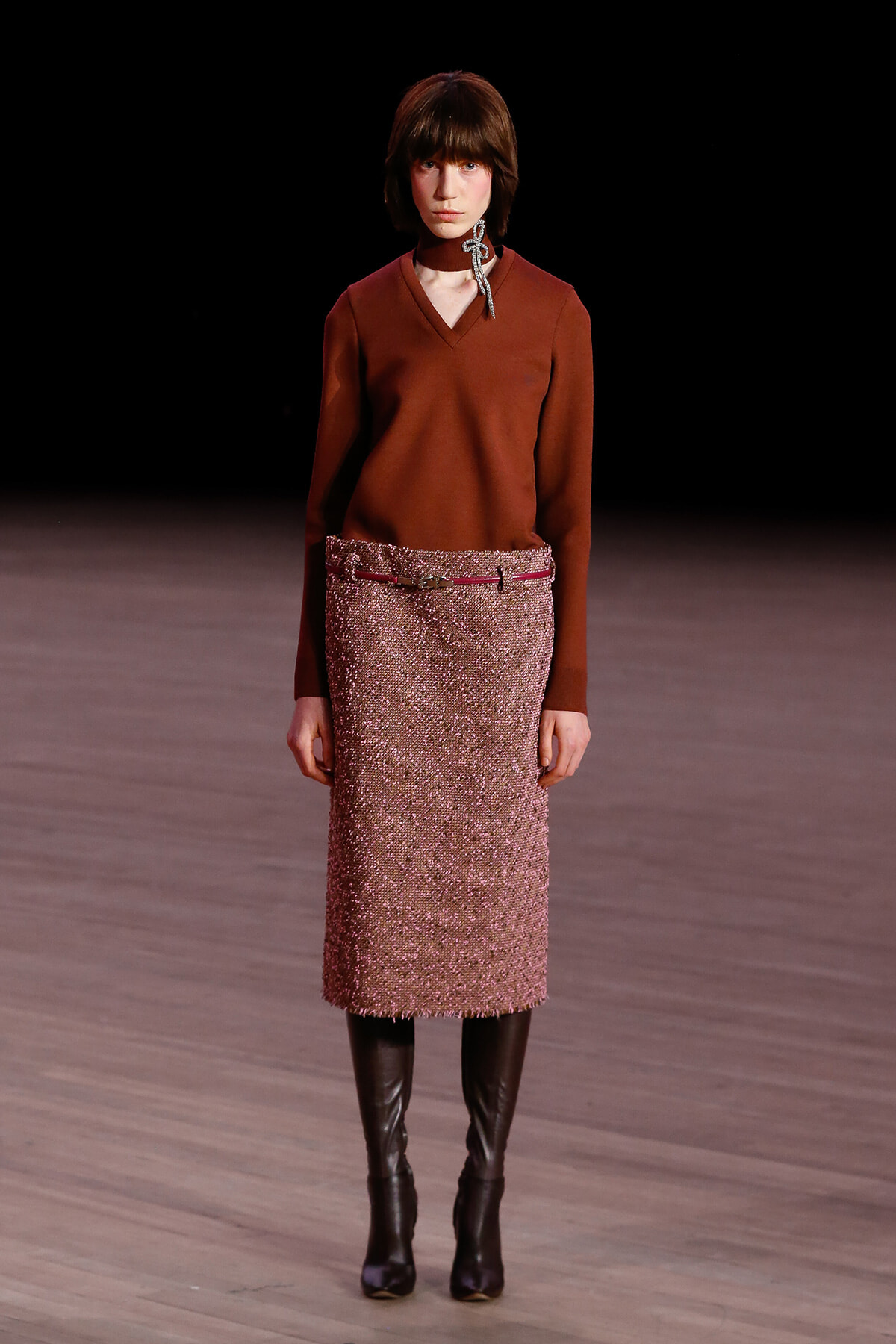 Female model walking a runway in a rust V-neck sweater, pink tweed knee-length skirt with a belt, and black knee-high boots, with a silver scarf tied at the neck.