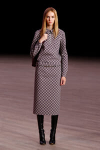 Model on a runway wearing a matching plaid midi dress with long sleeves, a zip collar, and a slim belt, paired with knee-high black boots against a dark background.